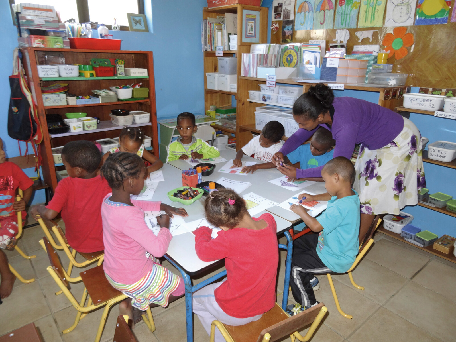 African children sit in a circle and paint. The teacher helps them with this. They are supported by a project called For Smiling Children, which is co-financed by the Grand Resort Bad Ragaz. The Grand Resort is a star hotel in Switzerland.