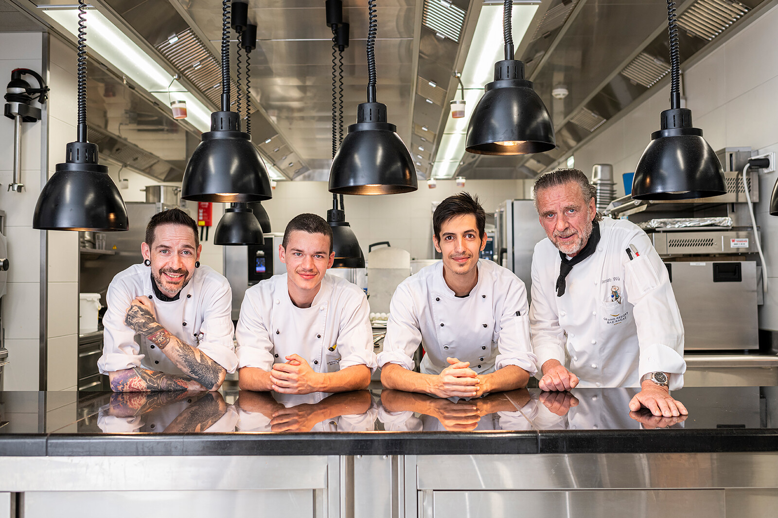 Four chefs, in their uniforms, lean on the kitchen counter. The kitchen can be seen in the background. The kitchen belongs to the Hotel Grand Resort Bad Ragaz.